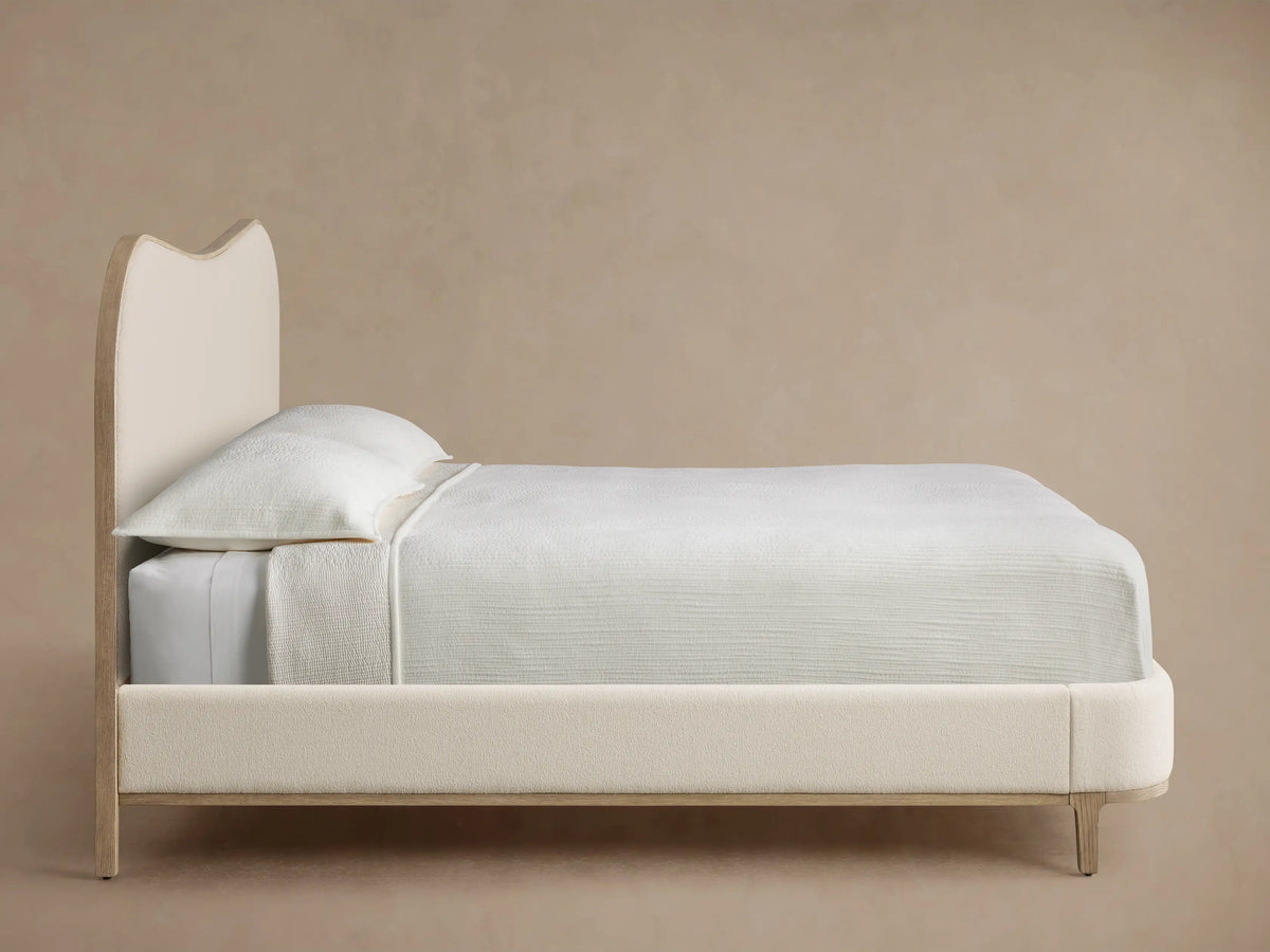 Close-up of Danny Camelback Bed headboard, showing curved light oak frame with wood grain and cream-colored textured upholstery.