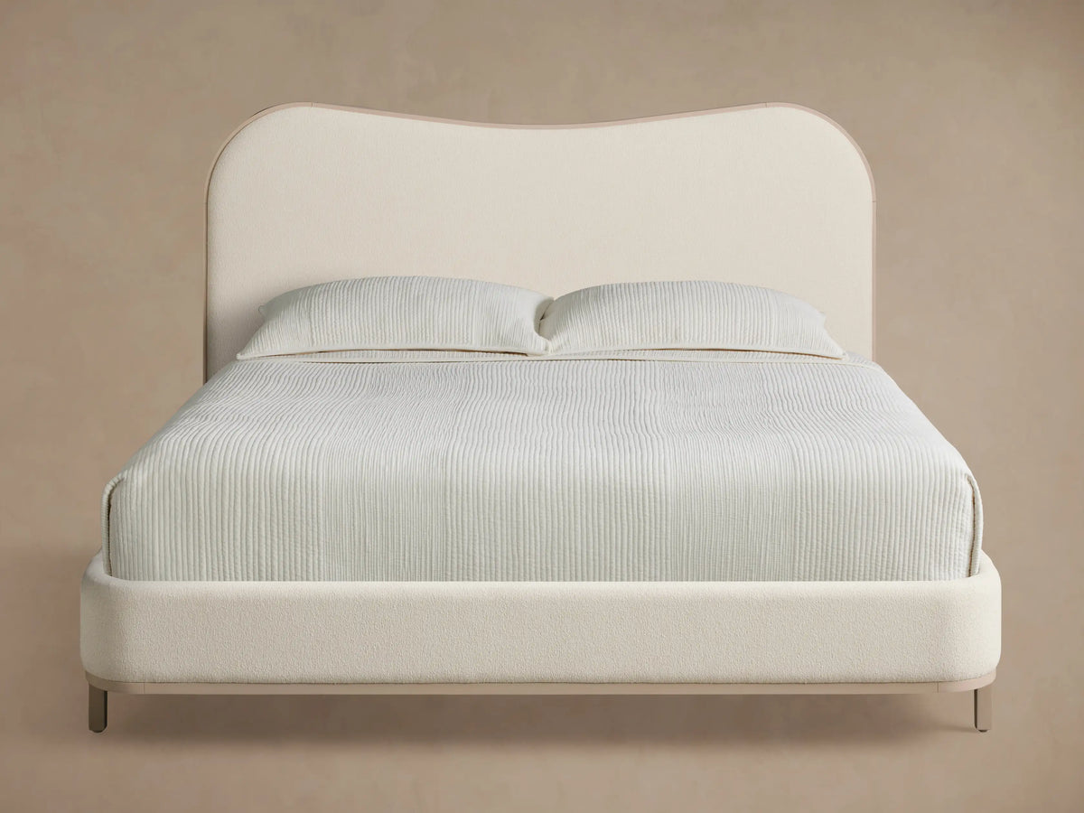 Danny Camelback Bed with a light beige upholstered camelback headboard & low frame, light grey legs. Striped bedding.