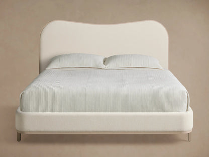 Danny Camelback Bed with a light beige upholstered camelback headboard & low frame, light grey legs. Striped bedding.