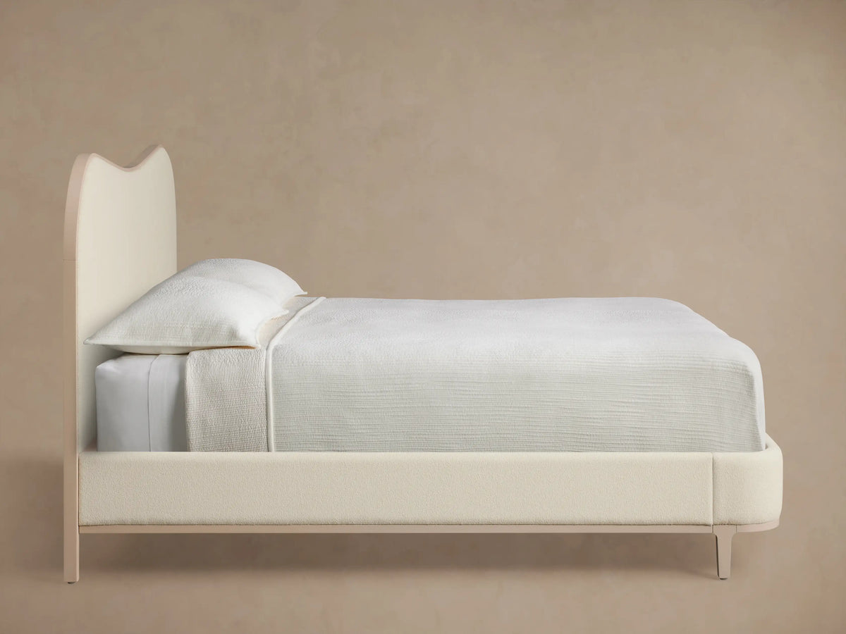 Close-up of Danny Camelback Bed's Fumo headboard. Curved corner, light cream textured upholstery, framed by smooth pale beige wood.