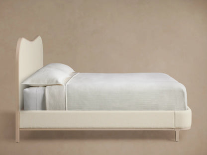 Close-up of Danny Camelback Bed's Fumo headboard. Curved corner, light cream textured upholstery, framed by smooth pale beige wood.