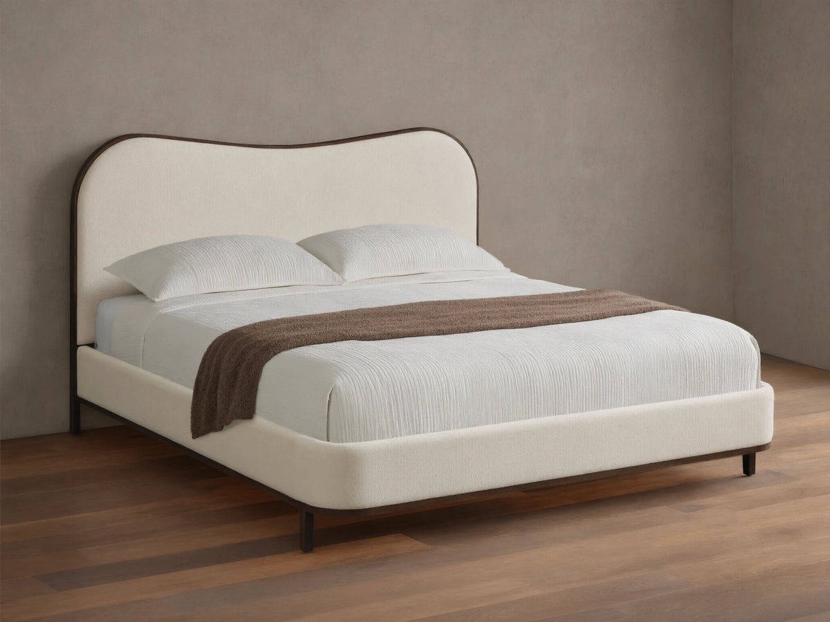 Cream upholstered Danny Camelback Bed with dark wood trim, light striped bedding, and a brown throw blanket.