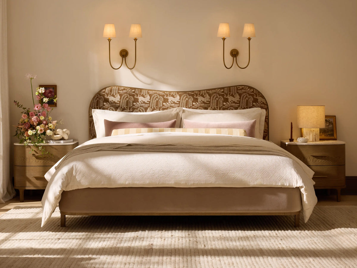 Cream upholstered Danny Camelback Bed with dark wood trim, light striped bedding, and a brown throw blanket.
