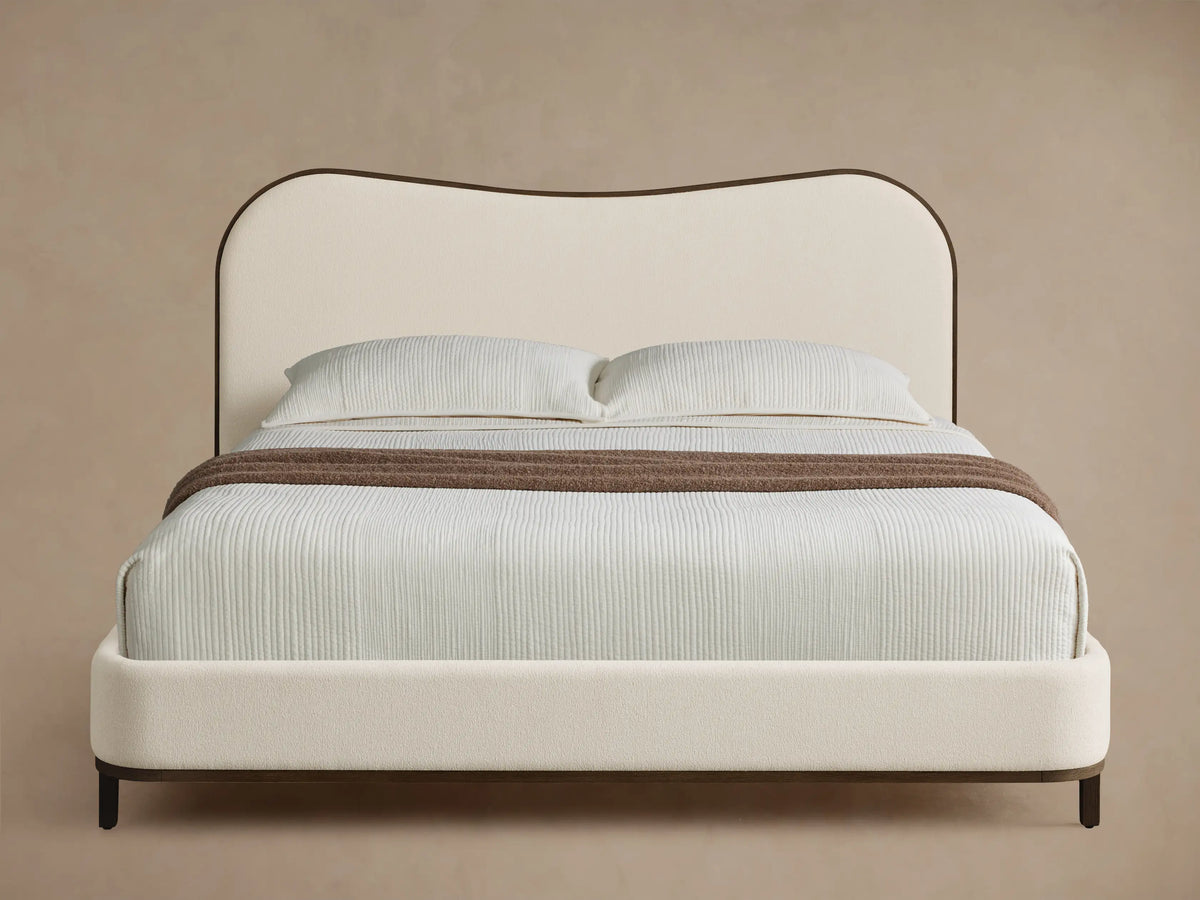 Front view of the Danny Camelback Bed. It has a brown patterned camelback headboard, a light brown upholstered base, and cream bedding.
