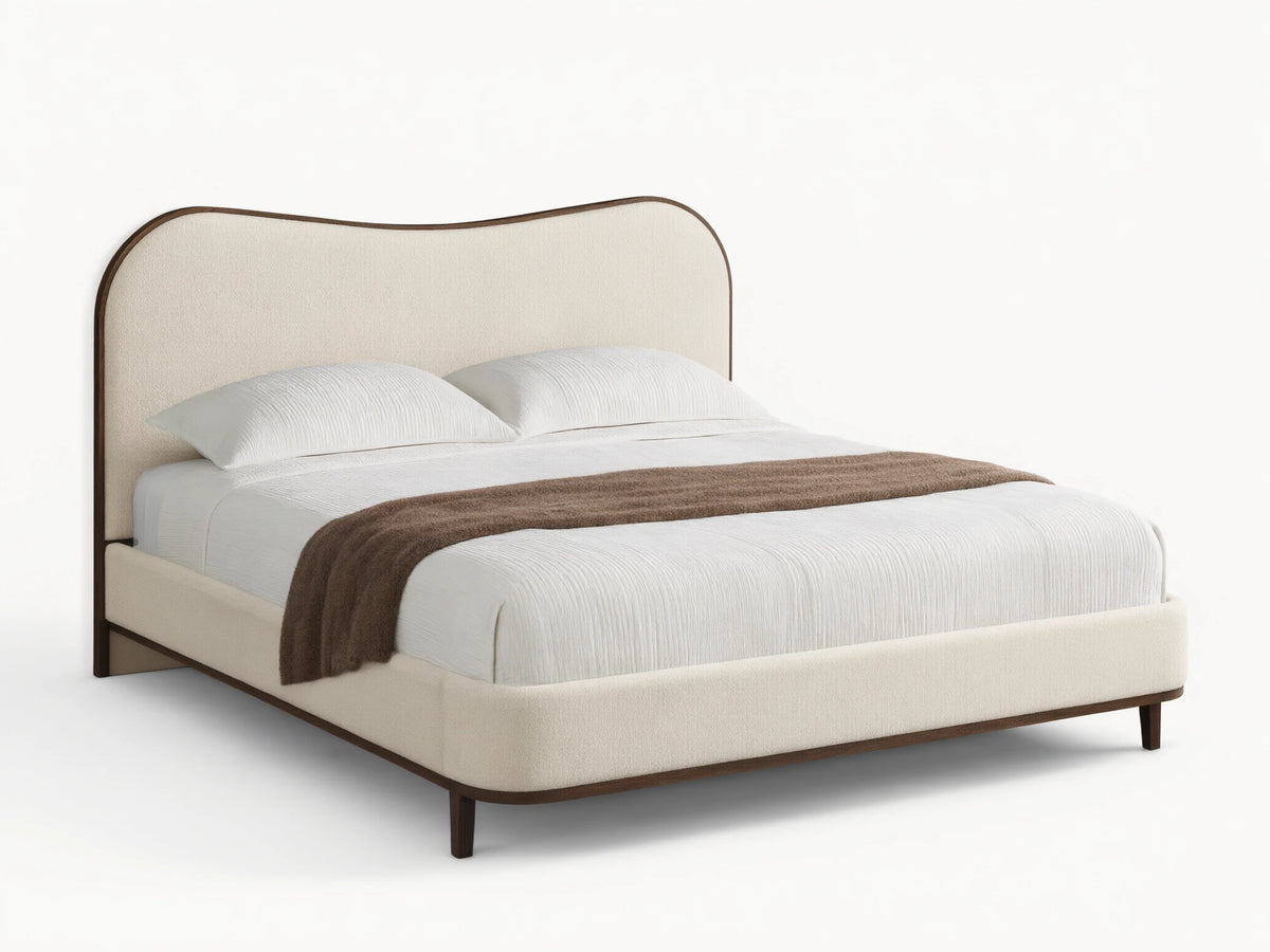 Cream upholstered Danny Camelback Bed with dark wood trim, light striped bedding, and a brown throw blanket.