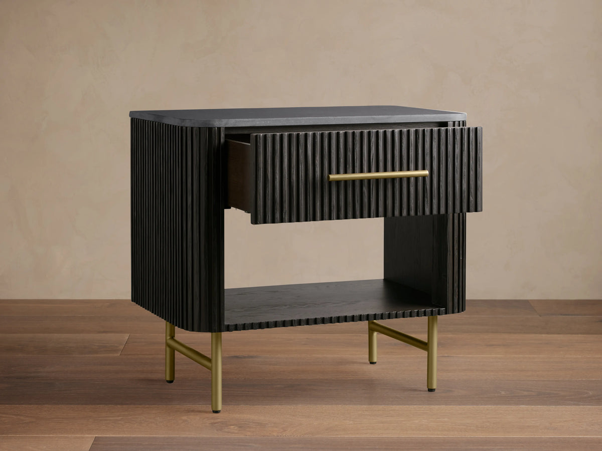 Finnley Open Nightstand in dark wood with vertical grooves, a brass handle drawer, and open shelf. Brass legs on wooden floor.