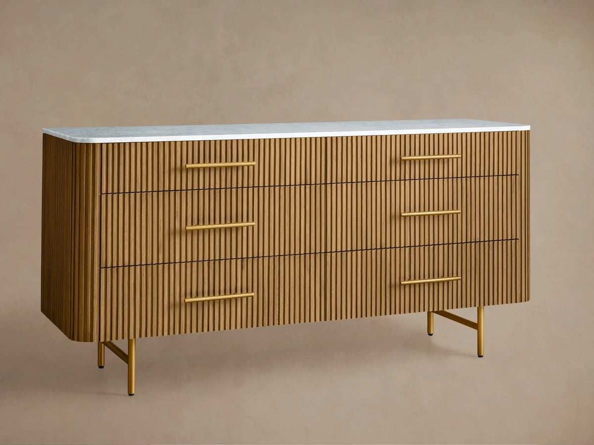 Finnley dresser with six honey-colored vertical slat drawers, gold handles, and Carrara white top, against a taupe wall.