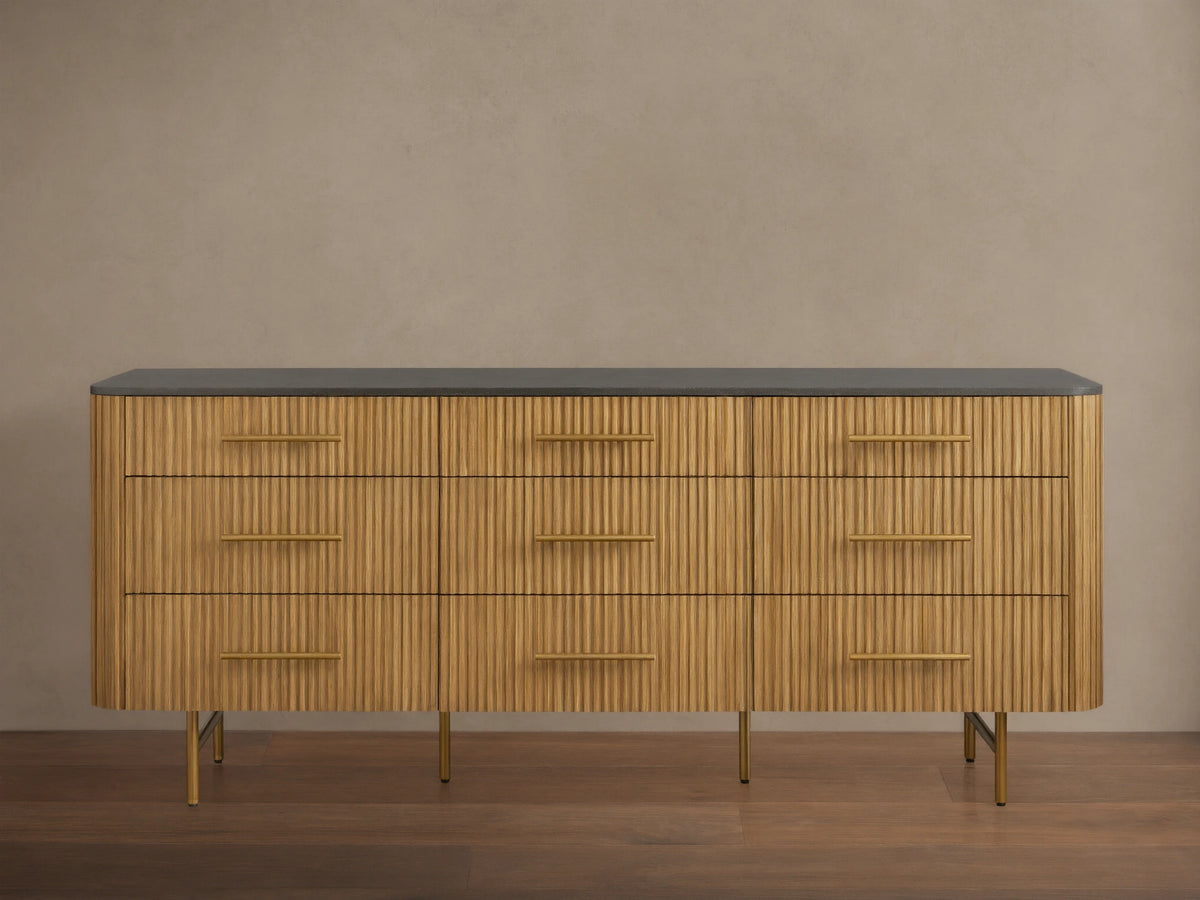 Front view of Finnley Nine-Drawer Dresser in honey-finished oak with vertical reeding, dark stone top, brass handles & legs.