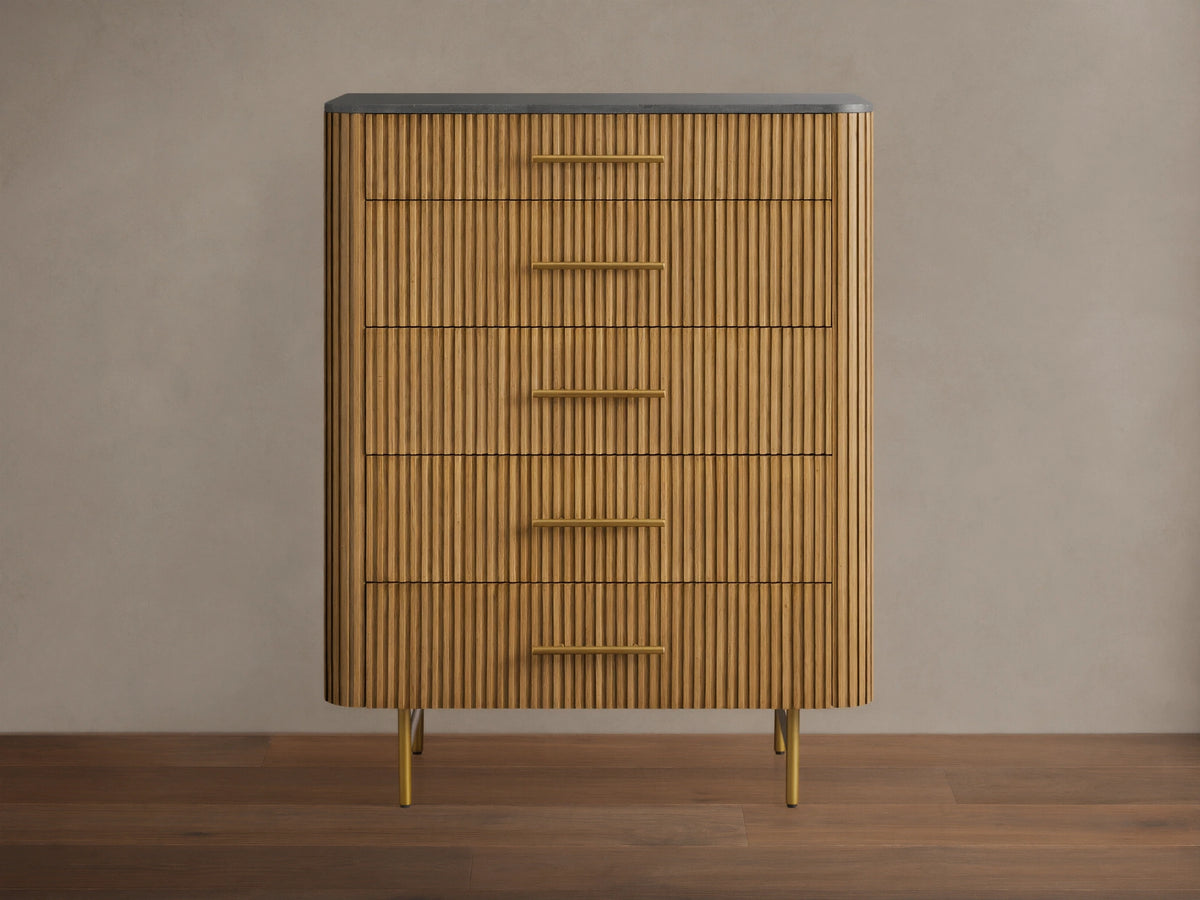 Finnley Five-Drawer Dresser. Honey wood with vertical reeding texture, five brass handles, dark stone top, and slim brass legs.