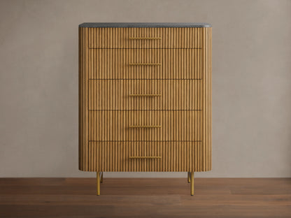 Finnley Five-Drawer Dresser. Honey wood with vertical reeding texture, five brass handles, dark stone top, and slim brass legs.