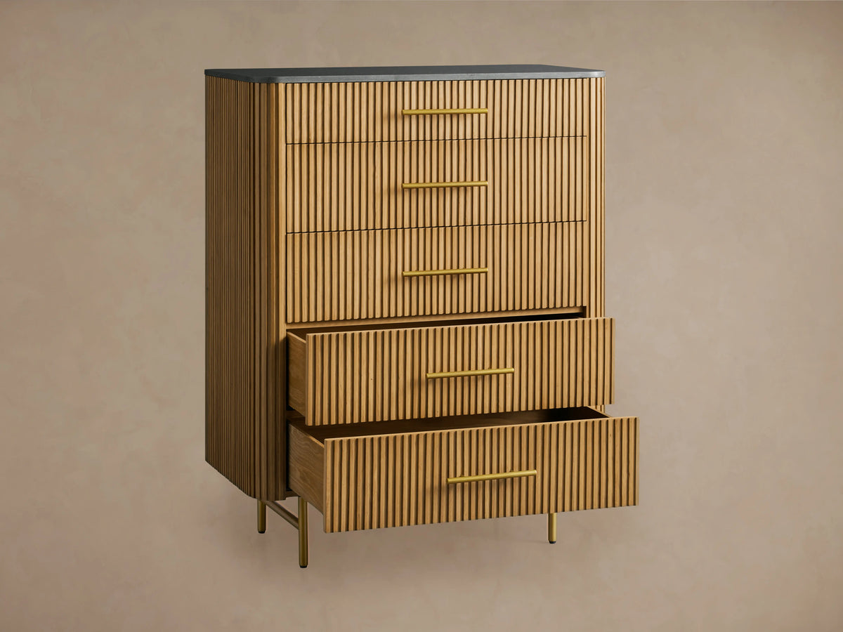 Finnley Five-Drawer Dresser. Honey wood with vertical reeding texture, five brass handles, dark stone top, and slim brass legs.