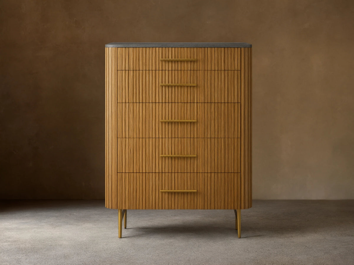 Finnley Five-Drawer Dresser. Honey wood with vertical reeding texture, five brass handles, dark stone top, and slim brass legs.
