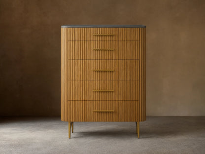 Finnley Five-Drawer Dresser. Honey wood with vertical reeding texture, five brass handles, dark stone top, and slim brass legs.