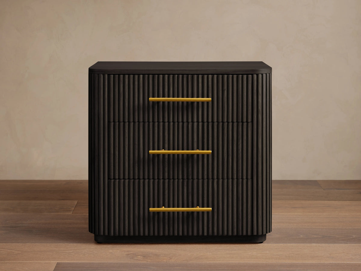 Finnley Wood Nightstand in Ebony with vertical grooves, featuring three drawers with brass finish handles, set on wooden floor.