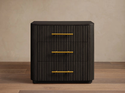 Finnley Wood Nightstand in Ebony with vertical grooves, featuring three drawers with brass finish handles, set on wooden floor.