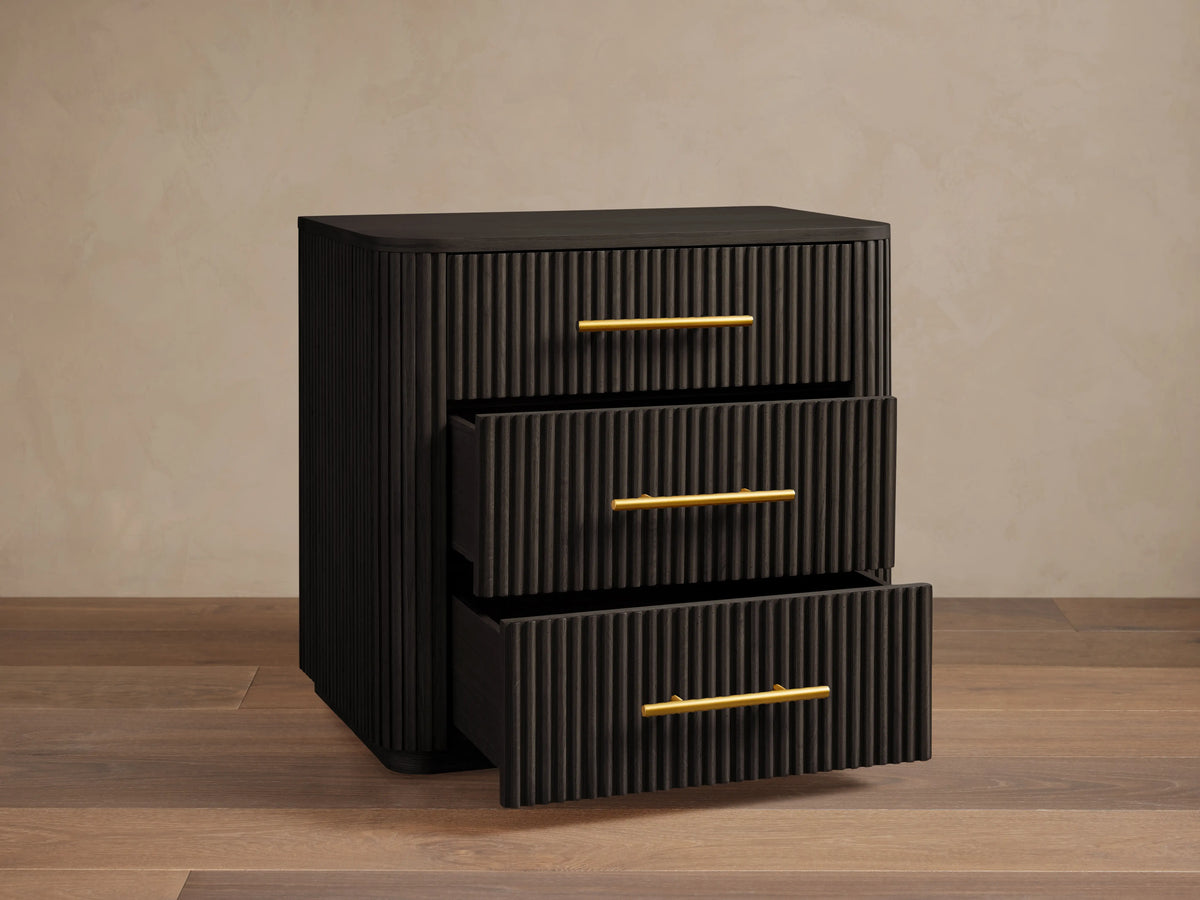 Finnley Wood Nightstand in Ebony with vertical grooves, featuring three drawers with brass finish handles, set on wooden floor.