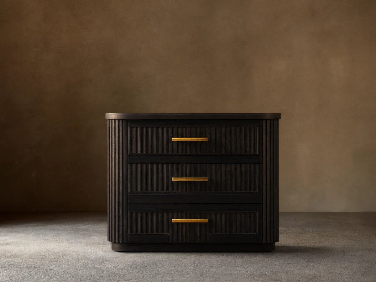 Finnley Wood Nightstand in Ebony with vertical grooves, featuring three drawers with brass finish handles, set on wooden floor.