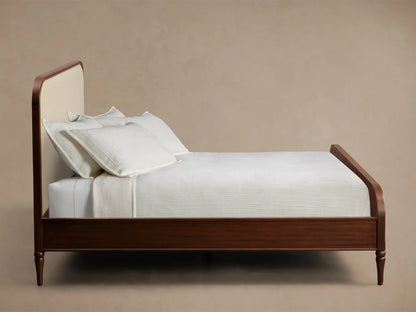 Side view of Frances Bed. Dark mahogany wood frame, rounded cream upholstered headboard, turned legs, white bedding.