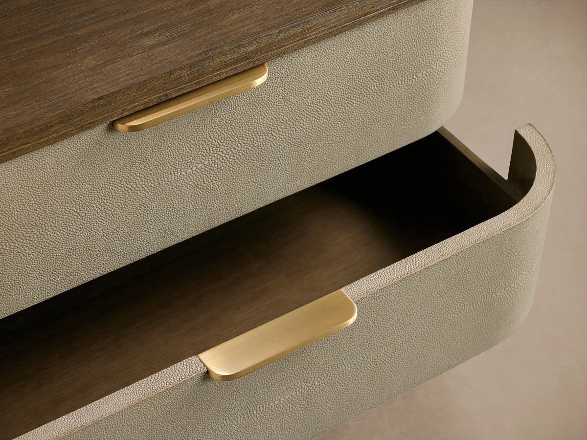Harrow Nightstand. Angled view of a 3-drawer nightstand with rounded light beige faux shagreen, dark wood top/base, brass pulls.