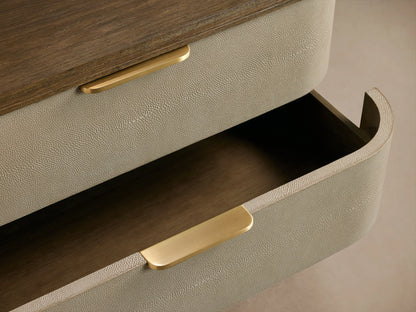Harrow Nightstand. Angled view of a 3-drawer nightstand with rounded light beige faux shagreen, dark wood top/base, brass pulls.