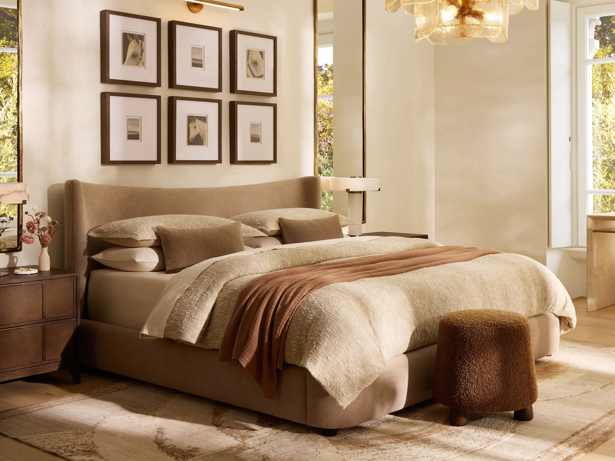 Hathaway Bed: Upholstered light brown wingback bed with cream bedding & rust throw blanket in a cozy room with art, a stool & side table.