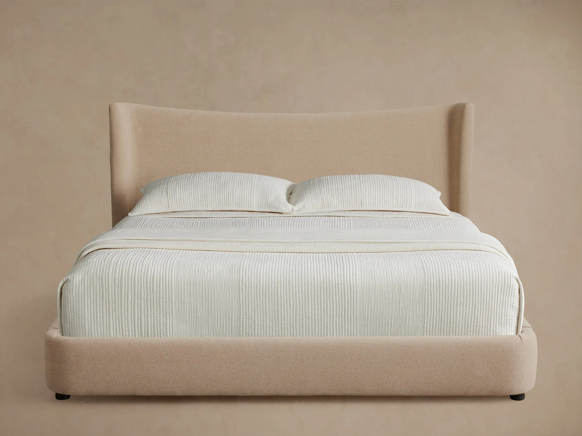 Front view of the beige upholstered Hathaway Bed, showing its curved headboard and platform, with off-white ribbed bedding.