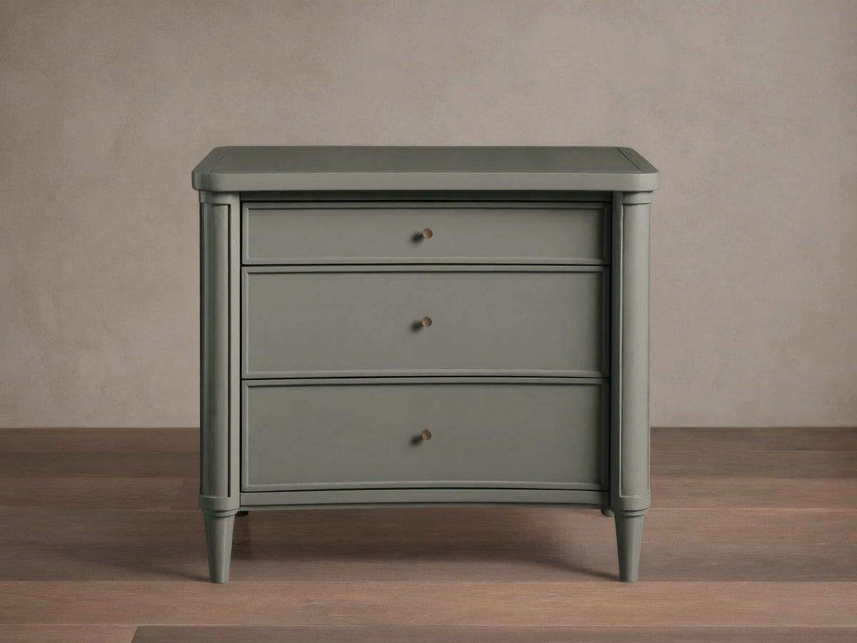 Henley Nightstand with three drawers, gray finish, petite brass pulls, and tapered legs on a wooden floor against a beige wall.