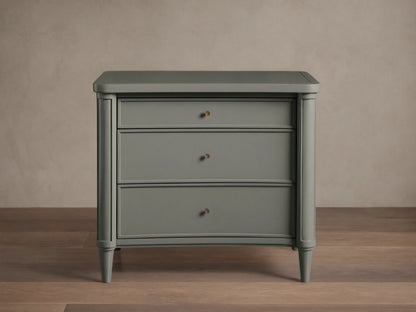 Henley Nightstand with three drawers, gray finish, petite brass pulls, and tapered legs on a wooden floor against a beige wall.