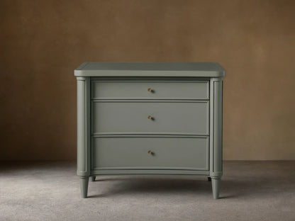 Henley Nightstand with three drawers, gray finish, petite brass pulls, and tapered legs on a wooden floor against a beige wall.