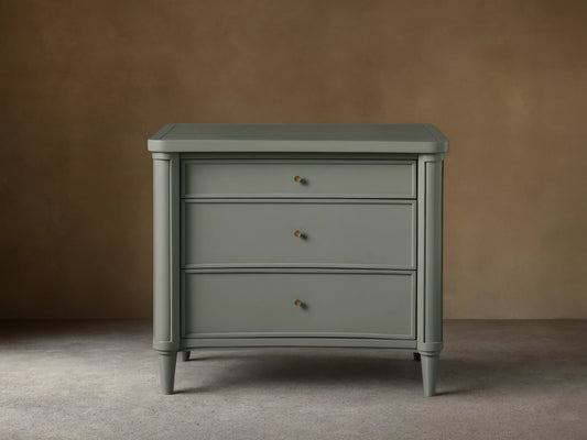 Henley Nightstand with three drawers, gray finish, petite brass pulls, and tapered legs on a wooden floor against a beige wall.