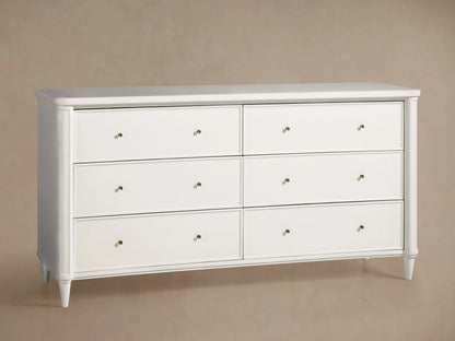 Front view of dark gray Henley Wide Six-Drawer Dresser with brass pulls, a lamp, and vase of flowers in a bedroom.