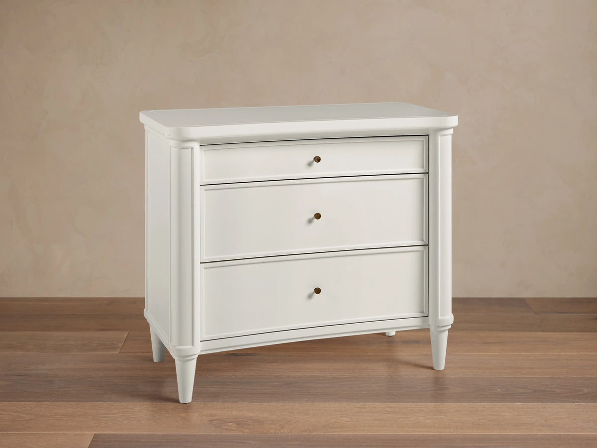 The Henley Nightstand is white with three drawers featuring petite brass pulls. It has rounded corners and tapered legs, set on a wooden floor.