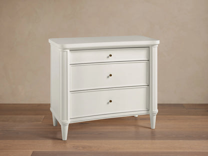 The Henley Nightstand is white with three drawers featuring petite brass pulls. It has rounded corners and tapered legs, set on a wooden floor.