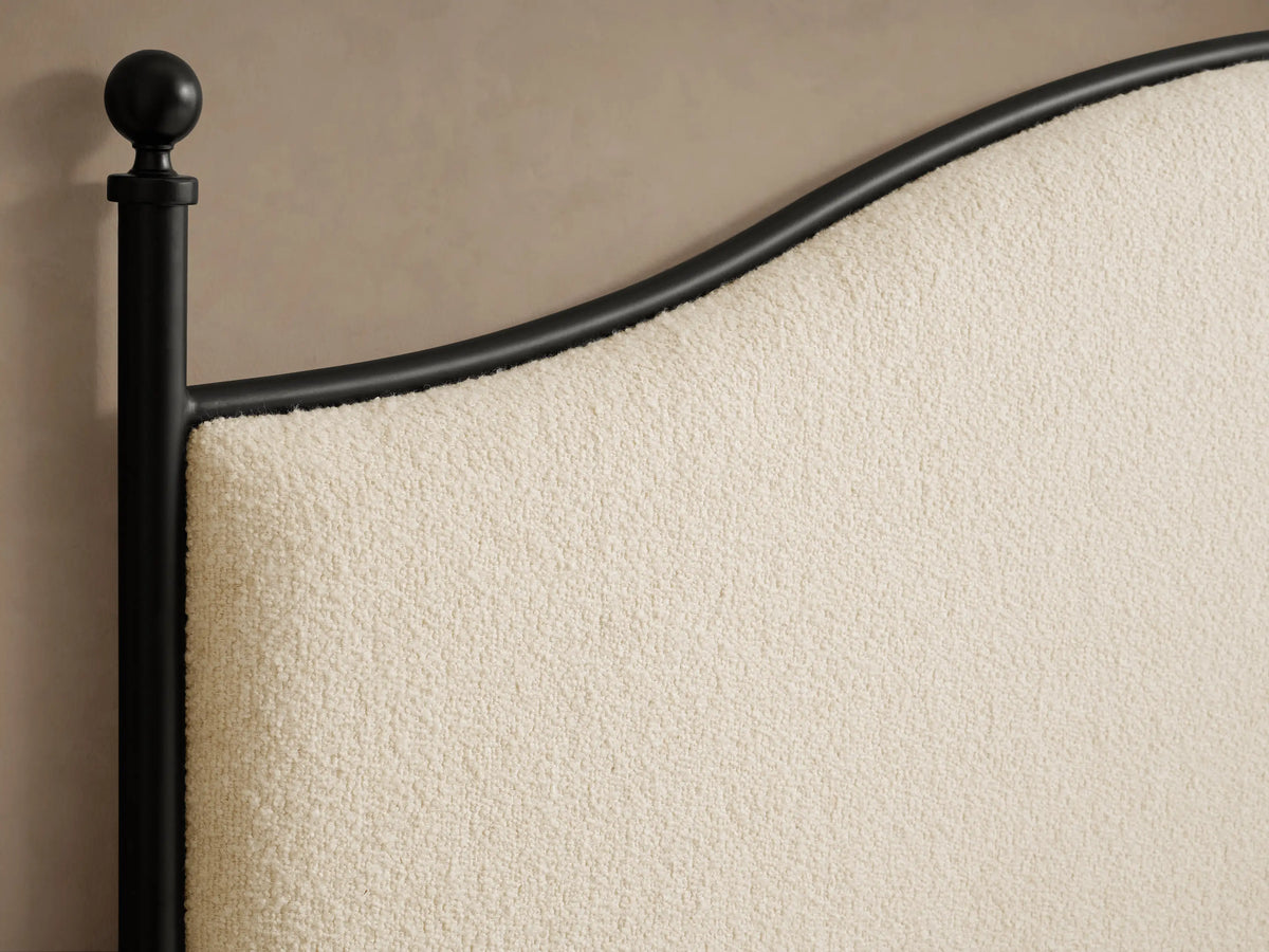 Side view of the Kimber Upholstered Bed with matte black iron frame, finial details, cream upholstered panels, and white bedding.