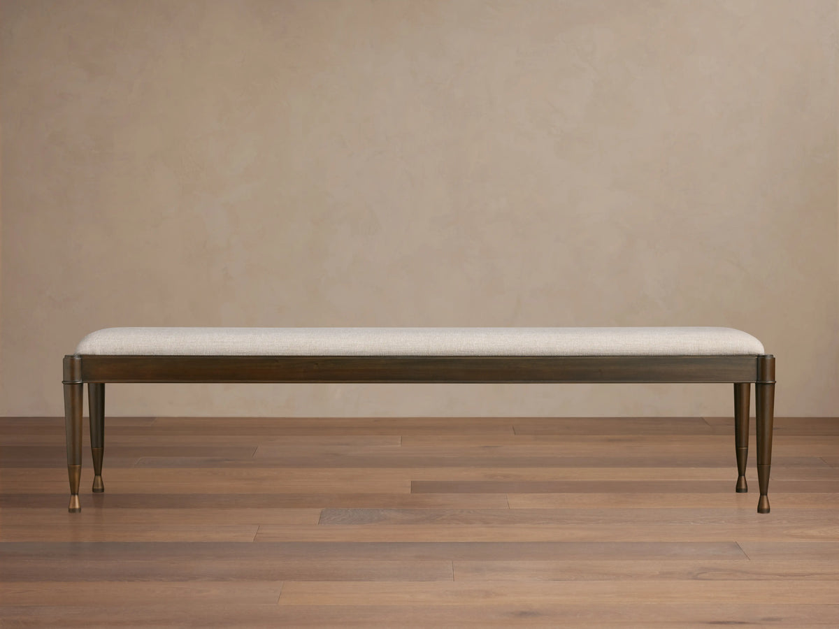 Laurent Bench with a long cushioned seat in light fabric, dark mahogany wood frame, and patinaed brass feet on wooden floor.