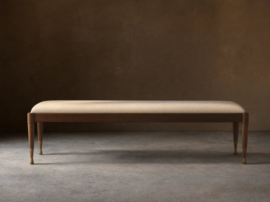 Laurent Bench with a long cushioned seat in light fabric, dark mahogany wood frame, and patinaed brass feet on wooden floor.