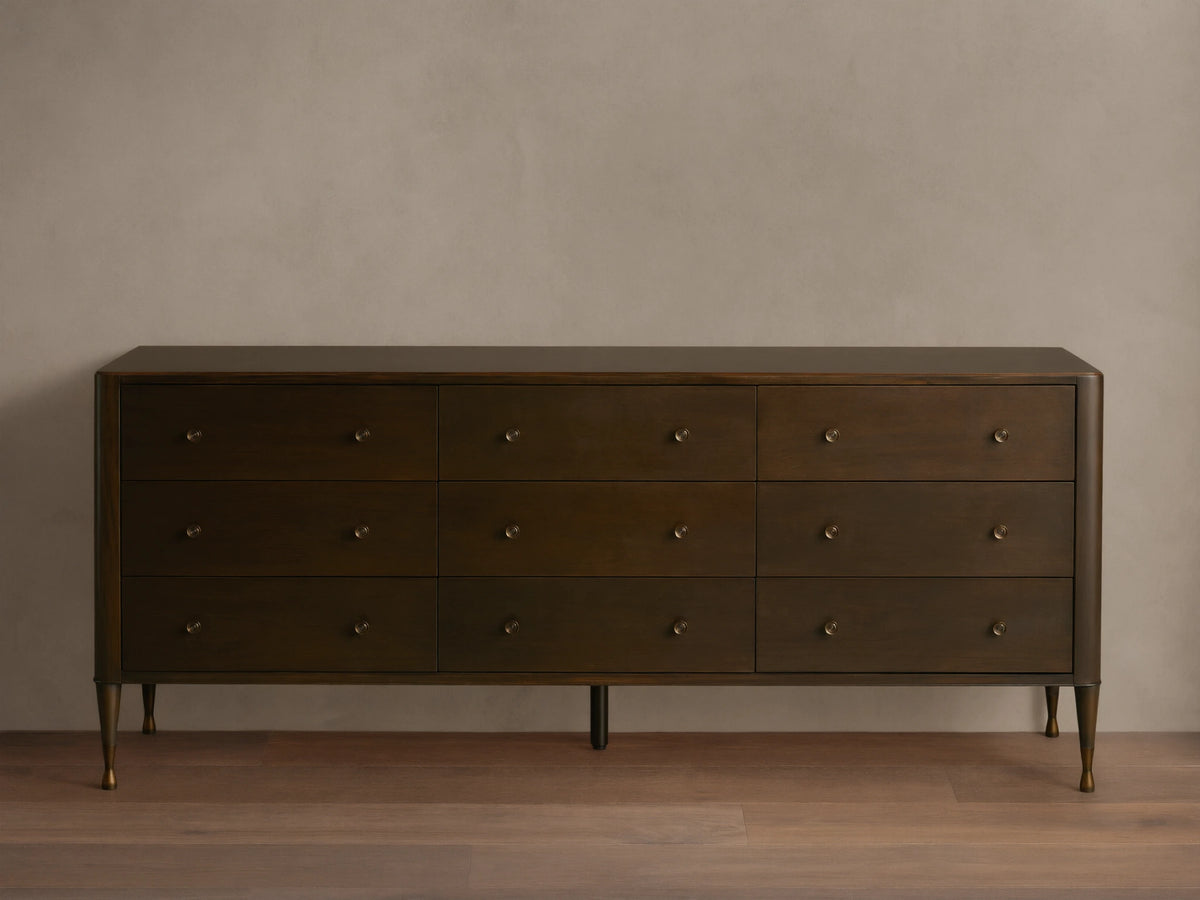 Long, dark brown Laurent Nine-Drawer Dresser with three rows of drawers, brass knobs, and tapered brass feet on a wooden floor.