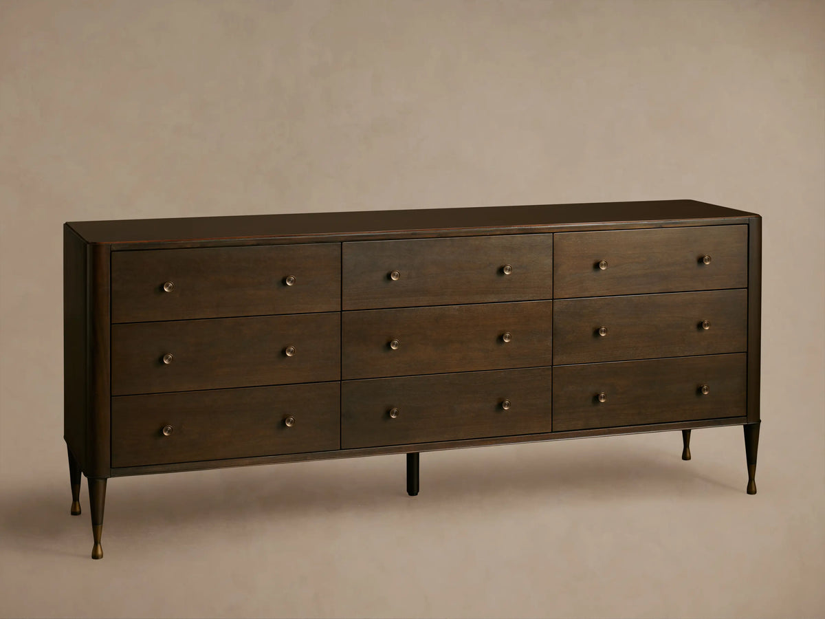 Long, dark brown Laurent Nine-Drawer Dresser with three rows of drawers, brass knobs, and tapered brass feet on a wooden floor.