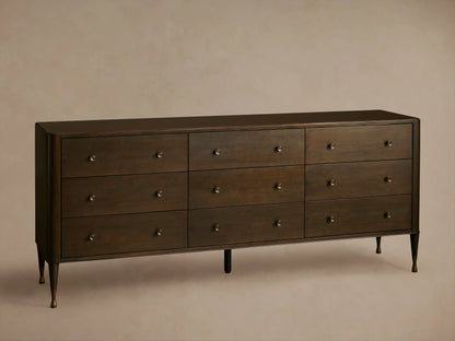 Long, dark brown Laurent Nine-Drawer Dresser with three rows of drawers, brass knobs, and tapered brass feet on a wooden floor.