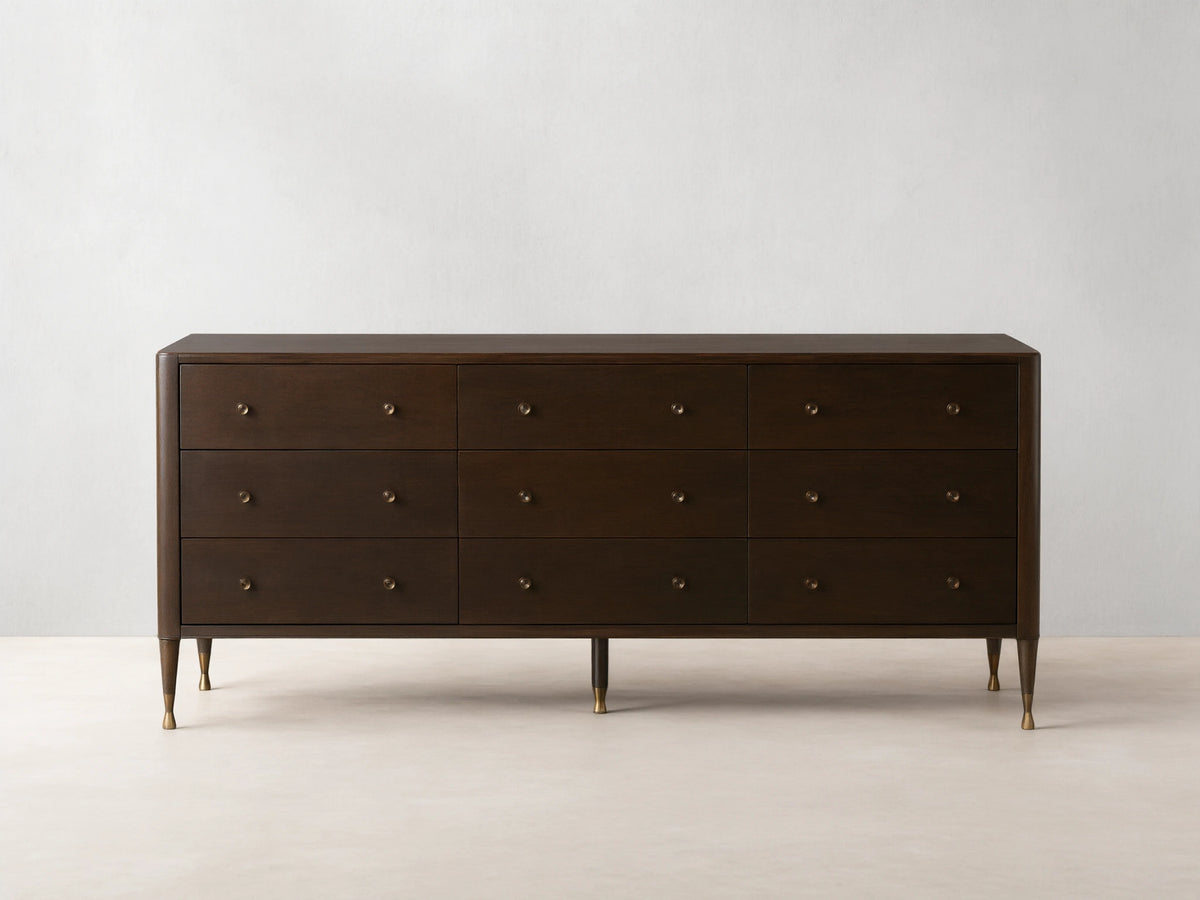 Long, dark brown Laurent Nine-Drawer Dresser with three rows of drawers, brass knobs, and tapered brass feet on a wooden floor.