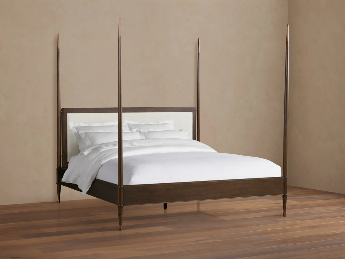 Four-poster Laurent Bed with mahogany wood frame and brass finials. White upholstered headboard, on wood floor in neutral room.
