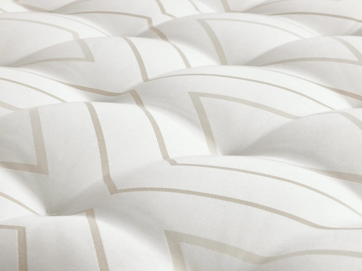 A luxury firm mattress on a light wood bed frame with a beige upholstered headboard. The white mattress features hand-tufting and two folded sheets on top.