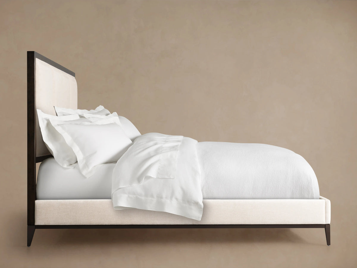 Malone Upholstered Bed, side view. Dark wood frame with bone upholstered headboard and footboard, topped with white bedding.