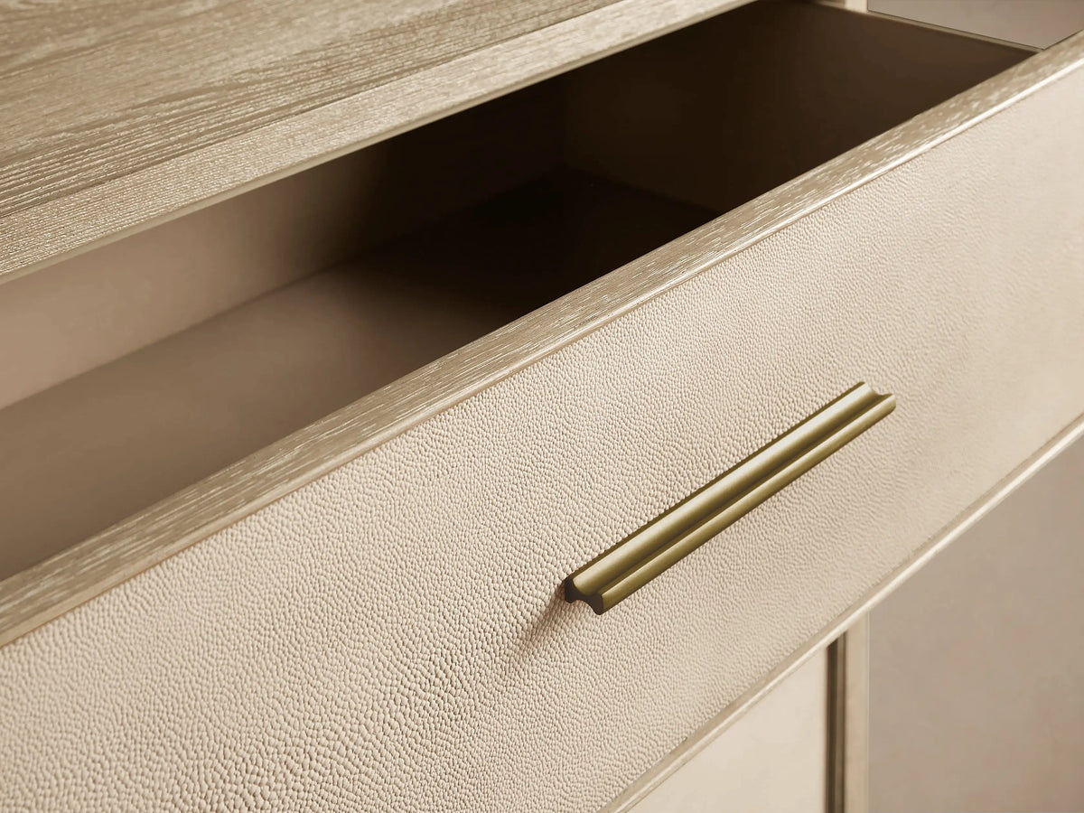 Angled view of Malone Three-Drawer Chest. Light wood frame, three cream shagreen-textured drawers, gold handles, metallic base.