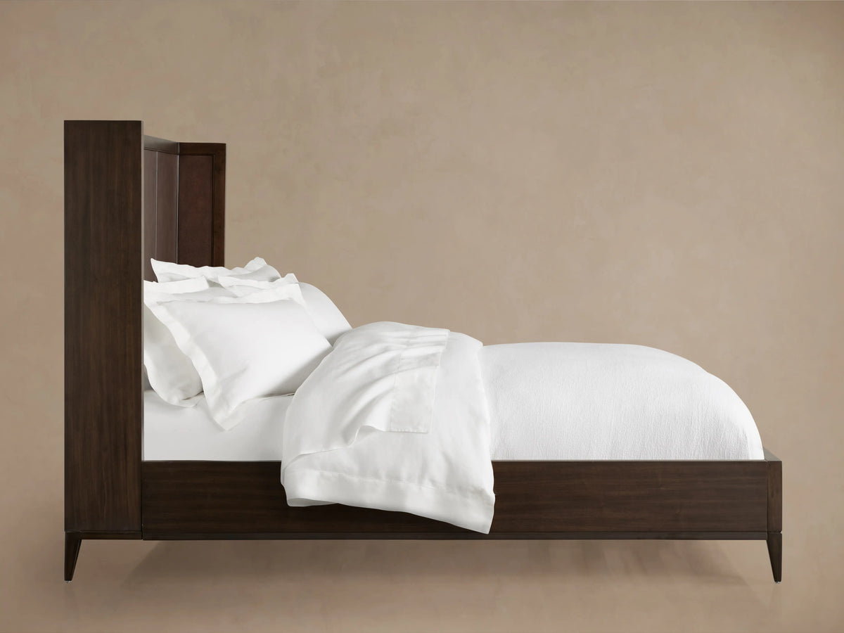 Malone Shelter Bed with dark wood frame and headboard, white bedding draped over the side, against a beige background.