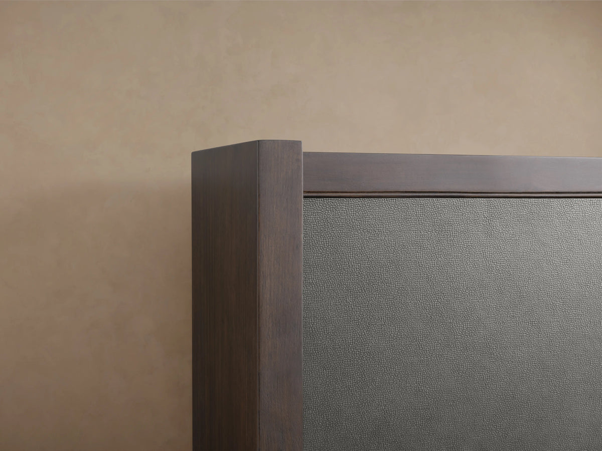 Angled close-up of Malone Shelter Bed corner, showing dark wood frame and dark grey textured leather panel against a beige wall.