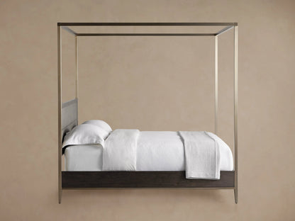 Malone Canopy Bed frame close-up: dark brown wood panel, brushed metallic leg, thin gold trim, white bedding above.