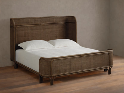 Maya Bed: Dark brown woven rattan bed frame with curved headboard & footboard, dressed with white sheets & pillows on a wood floor.