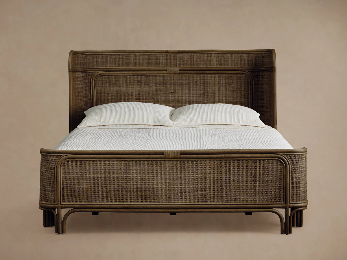 Maya Bed: Dark brown woven rattan bed frame with curved headboard & footboard, dressed with white sheets & pillows on a wood floor.