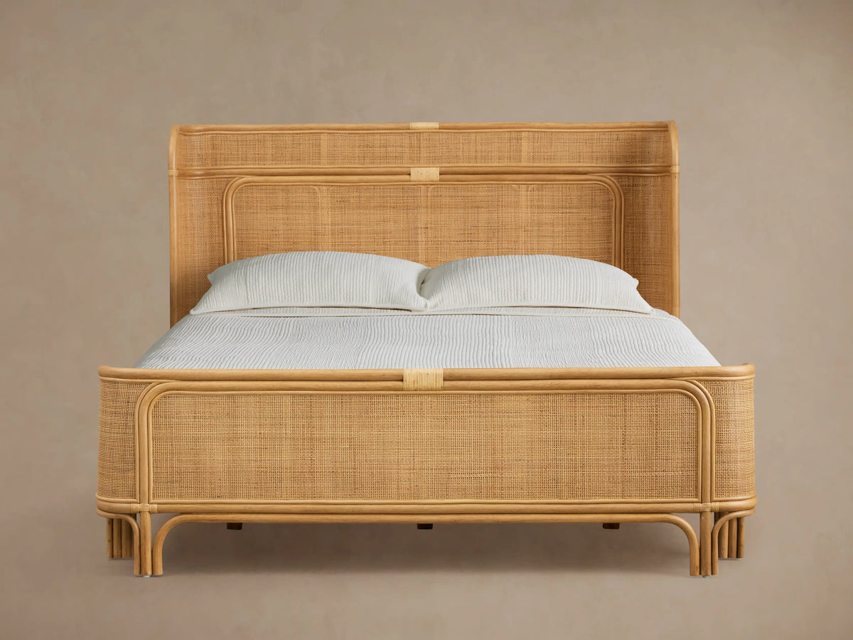 Natural rattan Maya Bed, with woven headboard & footboard, dressed with light blue bedding on a wooden floor.